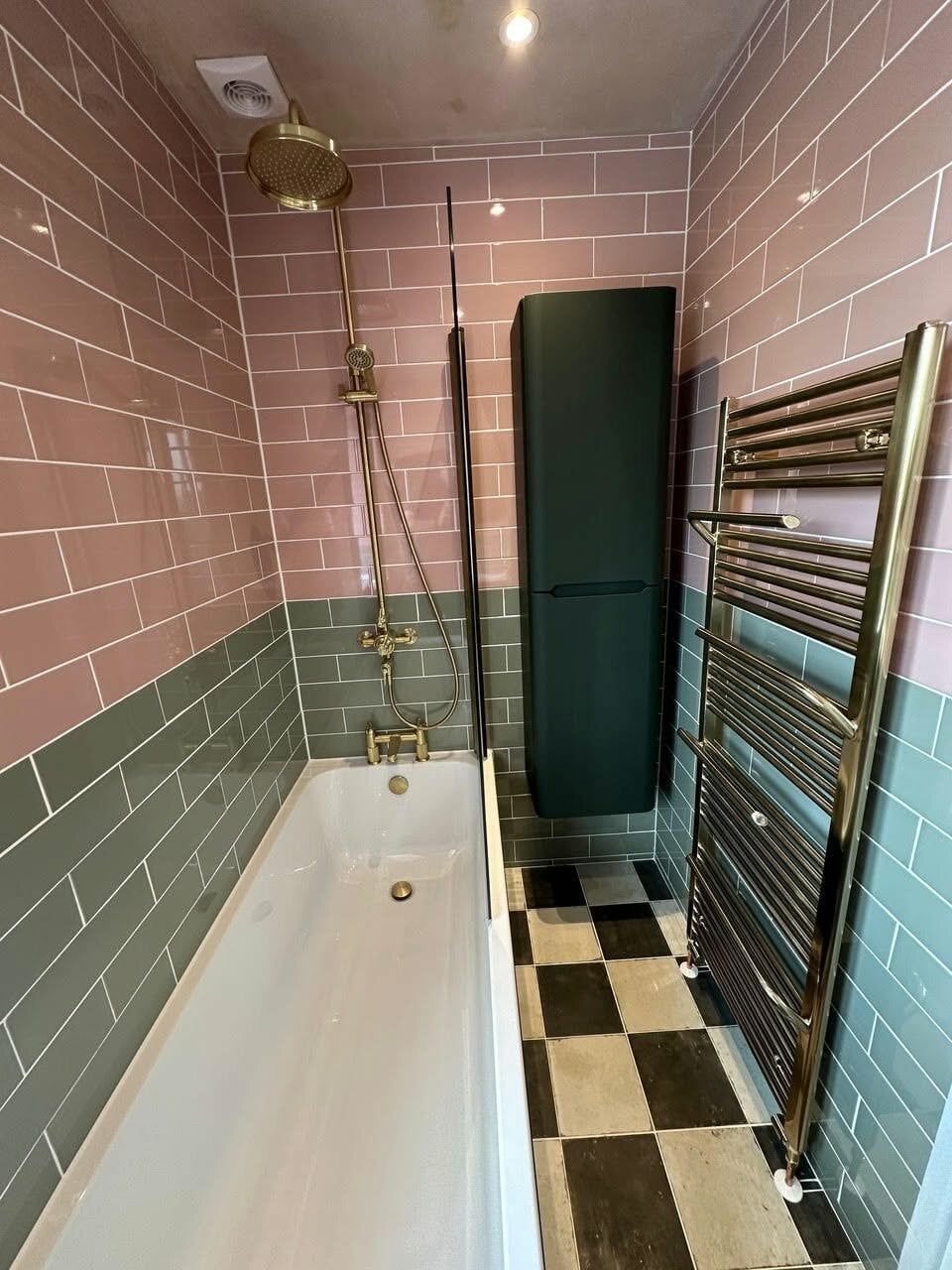 White bath surrounded by rose and sage gloss wall tiles with a forest green storage unit and brushed gold towel warmer.