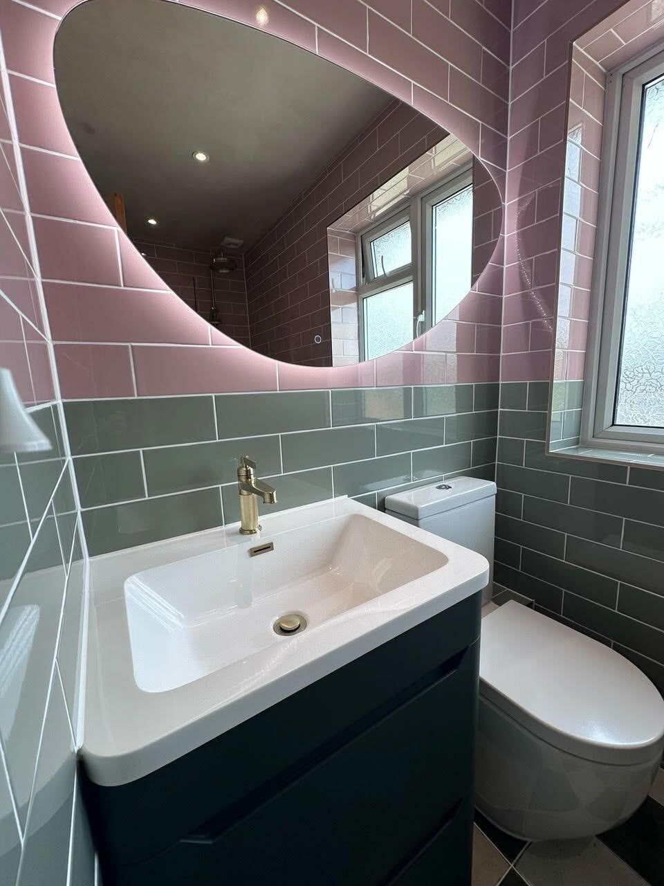 Forest green vanity unit with white basin, pebble-shaped mirror and brushed gold tap in a colourful tiled bathroom.