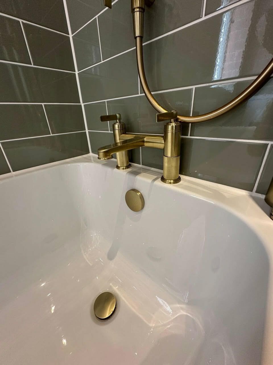 Close-up of a white basin with brushed gold tap against dark green gloss wall tiles and a pebble-shaped mirror.