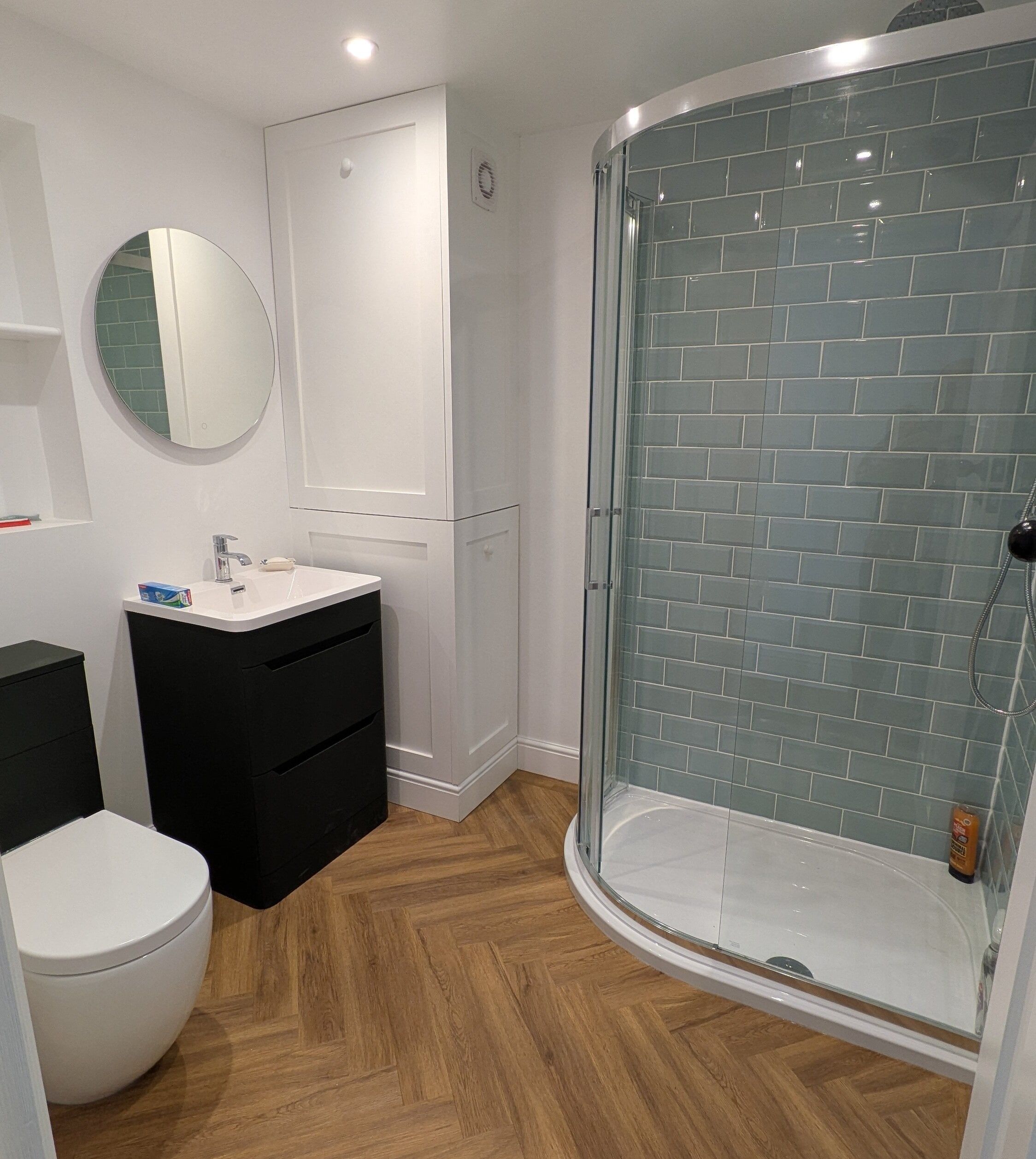 Customer bathroom renovation from Easy Bathrooms Tunbridge Wells featuring a dark vanity unit, curved shower enclosure, white sanitaryware and warm wood-effect flooring.