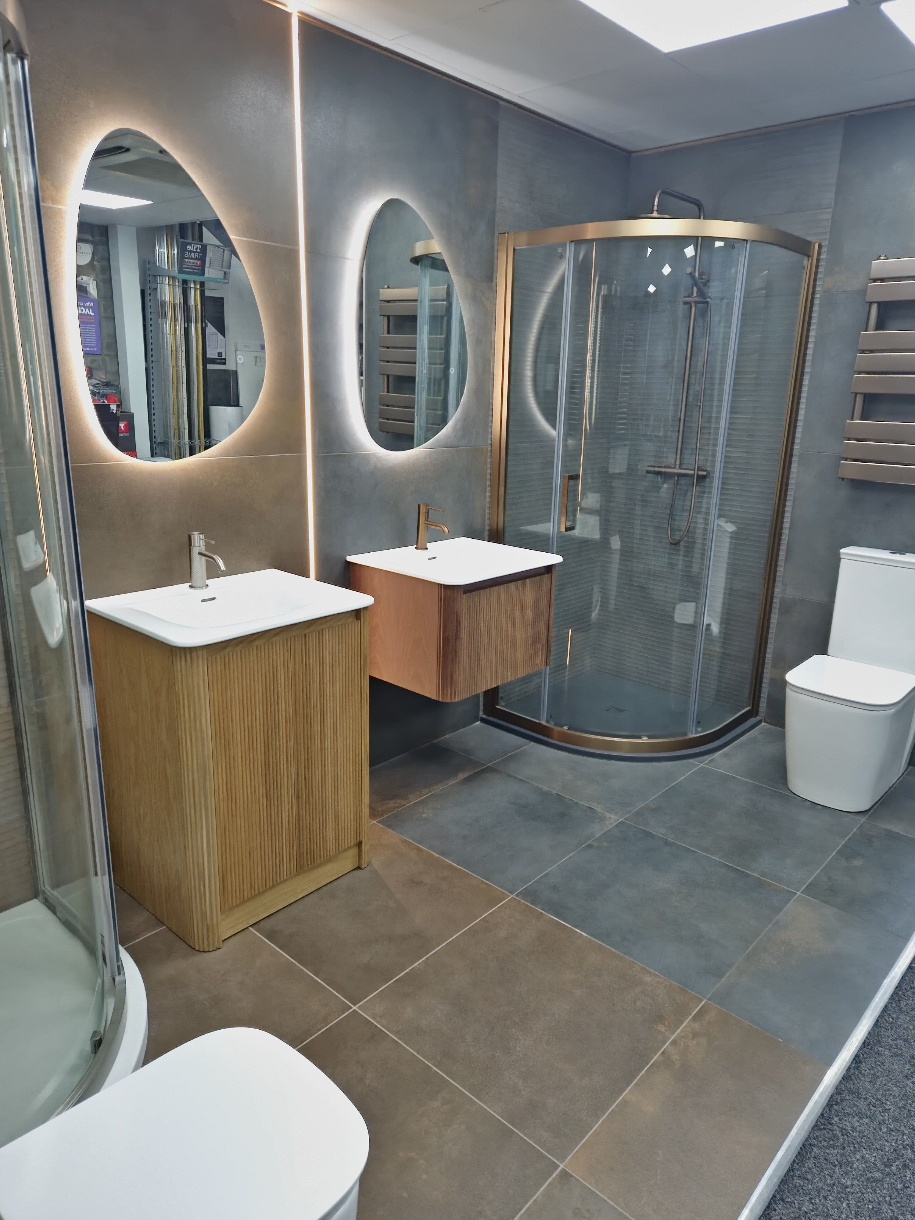 Contemporary bathroom display at Easy Bathrooms Basildon featuring wood-effect Wish furniture, illuminated mirrors, a curved shower enclosure and coordinating brassware.