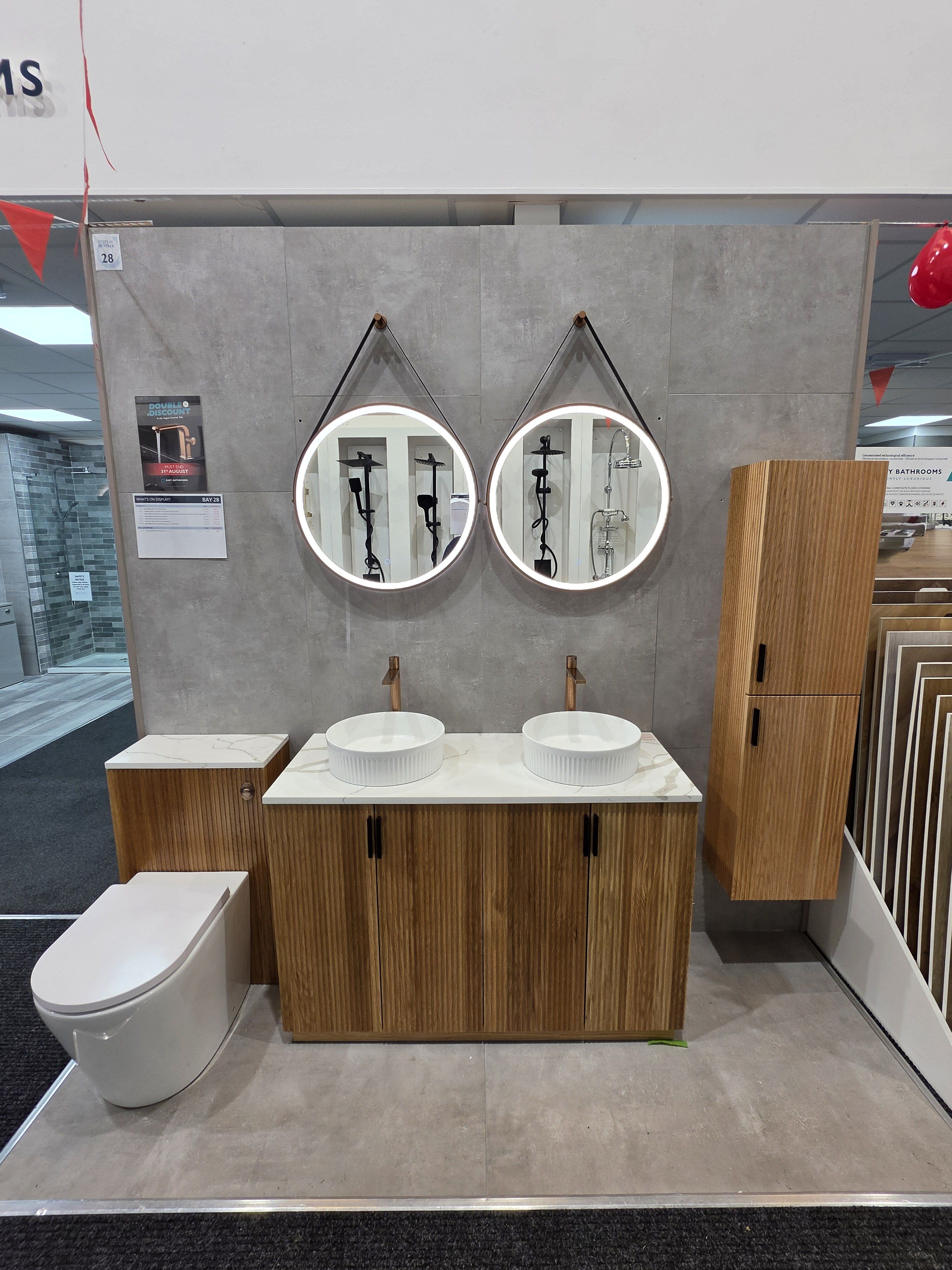 New bathroom display at Easy Bathrooms Chichester featuring fluted wood-effect furniture, twin countertop basins, brushed bronze fittings, illuminated mirrors and stone-effect tiles.