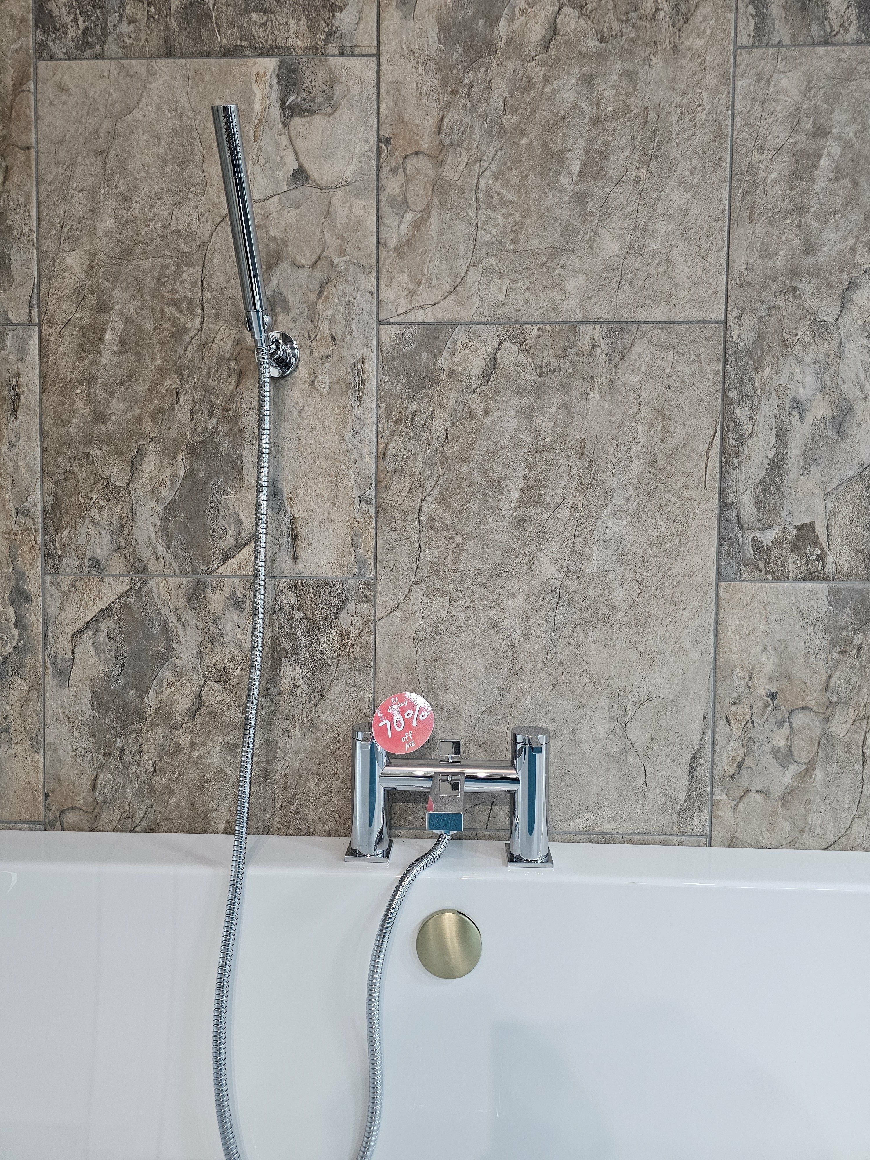 Chrome ex-display bath shower mixer and handset fitted against neutral stone-effect wall tiles.