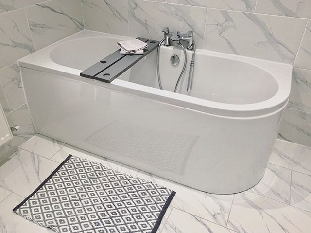 A modern white D-shaped back-to-wall bath with a chrome mixer tap, set against elegant grey-veined marble effect floor and wall tiles.
