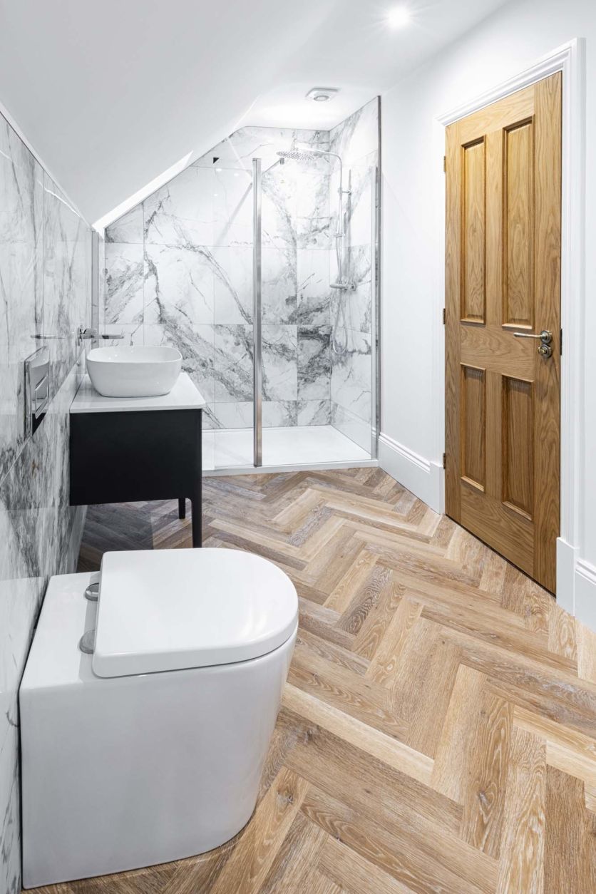 Modern bathroom with marble wall tiles, warm wood herringbone flooring, walk-in shower and grey floating vanity.