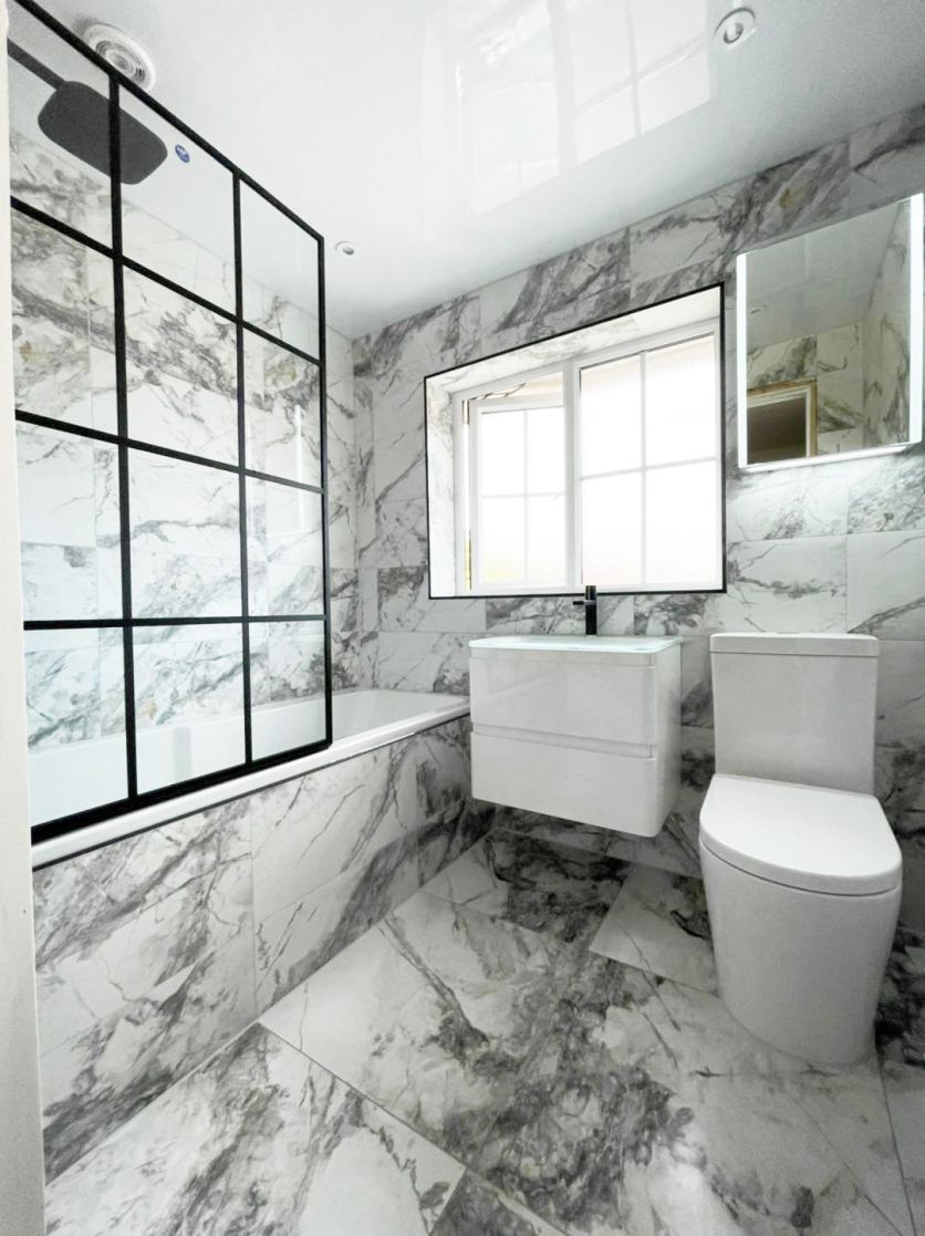 A striking modern bathroom featuring large-format grey and white marble effect tiles on the walls and floor, complemented by a black-framed Crittall-style shower screen and a sleek white wall-hung vanity unit with a minimalist black basin mixer tap.