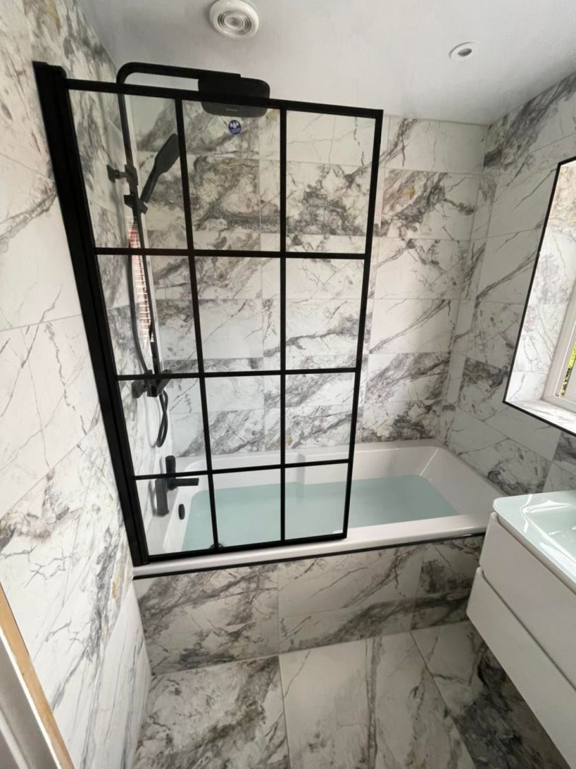 A contemporary shower bath setup featuring a black framed crittall-style glass screen and matching matt black rainfall shower head, set against floor-to-ceiling white marble effect tiles with deep grey veining.