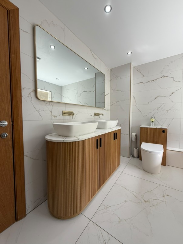 A large Revel ribbed natural oak double vanity with a marble worktop and Grain matt white stone basins, featuring Moray champagne gold wall-mounted taps and matching oak WC unit.