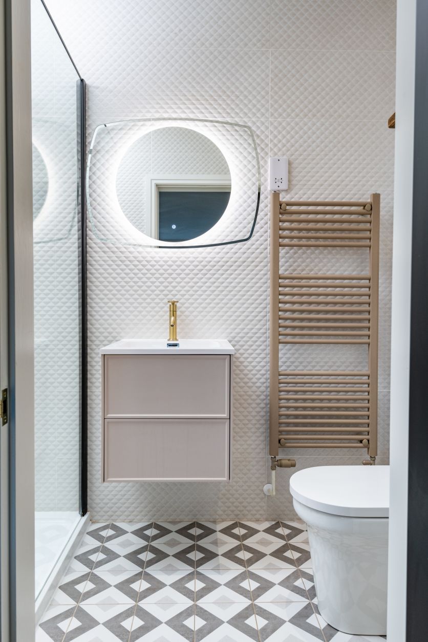 Modern blush and gold bathroom with textured white tiles, patterned floor, wall-hung vanity and LED backlit mirror.