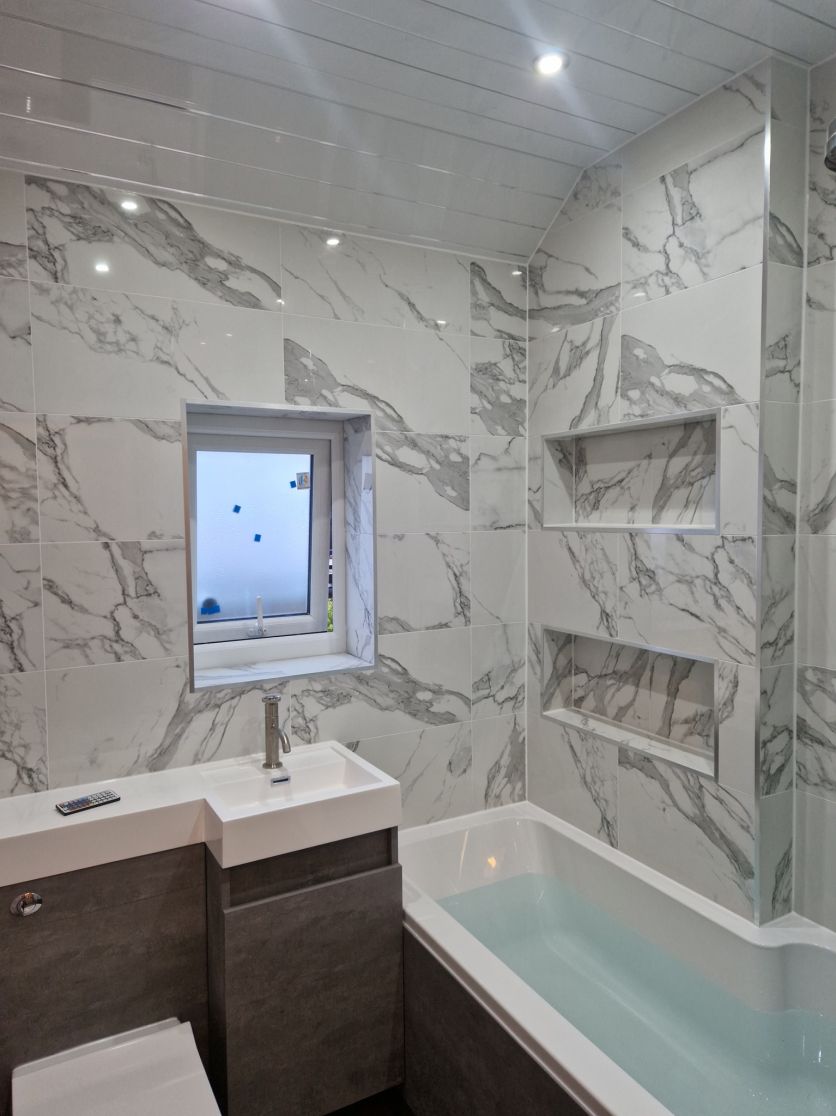 A contemporary small bathroom featuring floor-to-ceiling white marble effect wall tiles, a compact grey wood-effect vanity unit with an integrated white basin