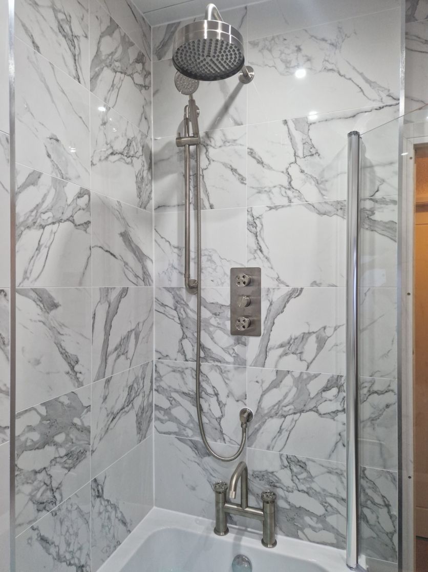 A contemporary shower-over-bath featuring large-format white marble effect wall tiles with deep grey veining, equipped with a luxury brushed steel rainfall shower head and a matching handheld spray.