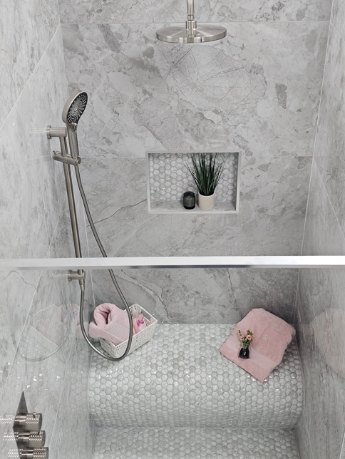 Walk-in shower display with grey marble-effect wall tiles, pebble mosaic seating, recessed storage niche and brushed metal shower fittings.