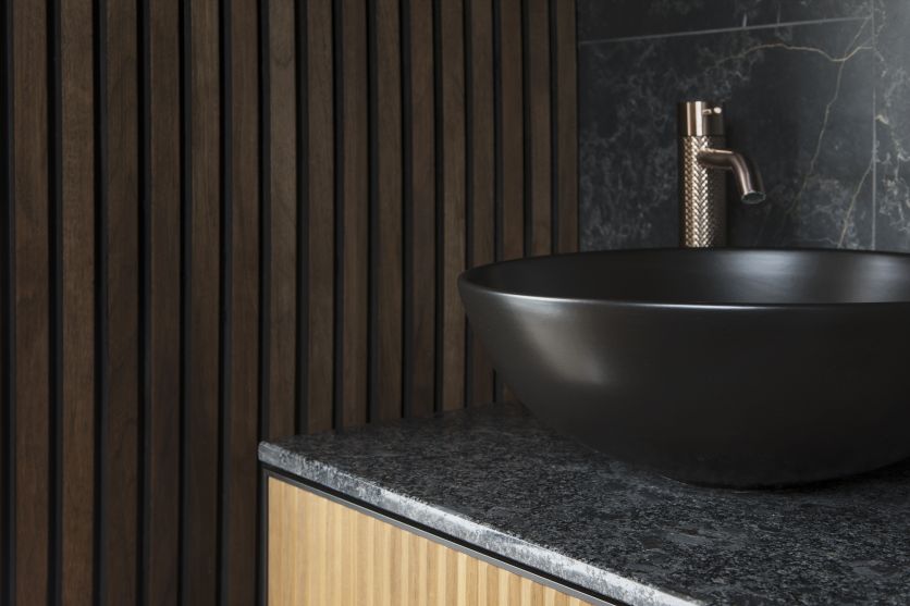 A matt black countertop vessel basin sits on a dark granite-style surface, paired with a knurled brushed brass mixer tap against a backdrop of dark wood slat wall panelling and black marble tiles.