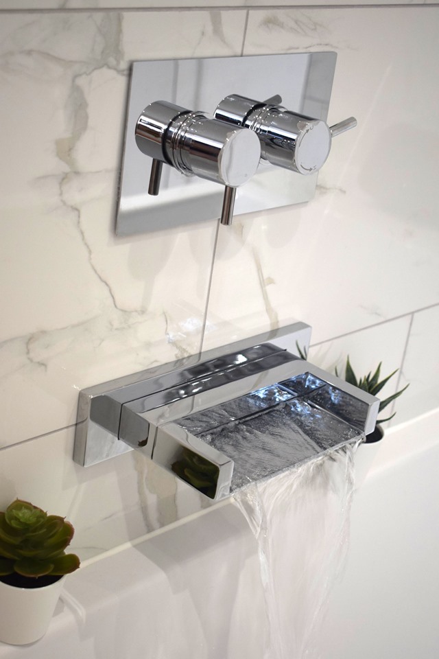 A contemporary wall-mounted chrome waterfall bath tap flowing into a white tub.