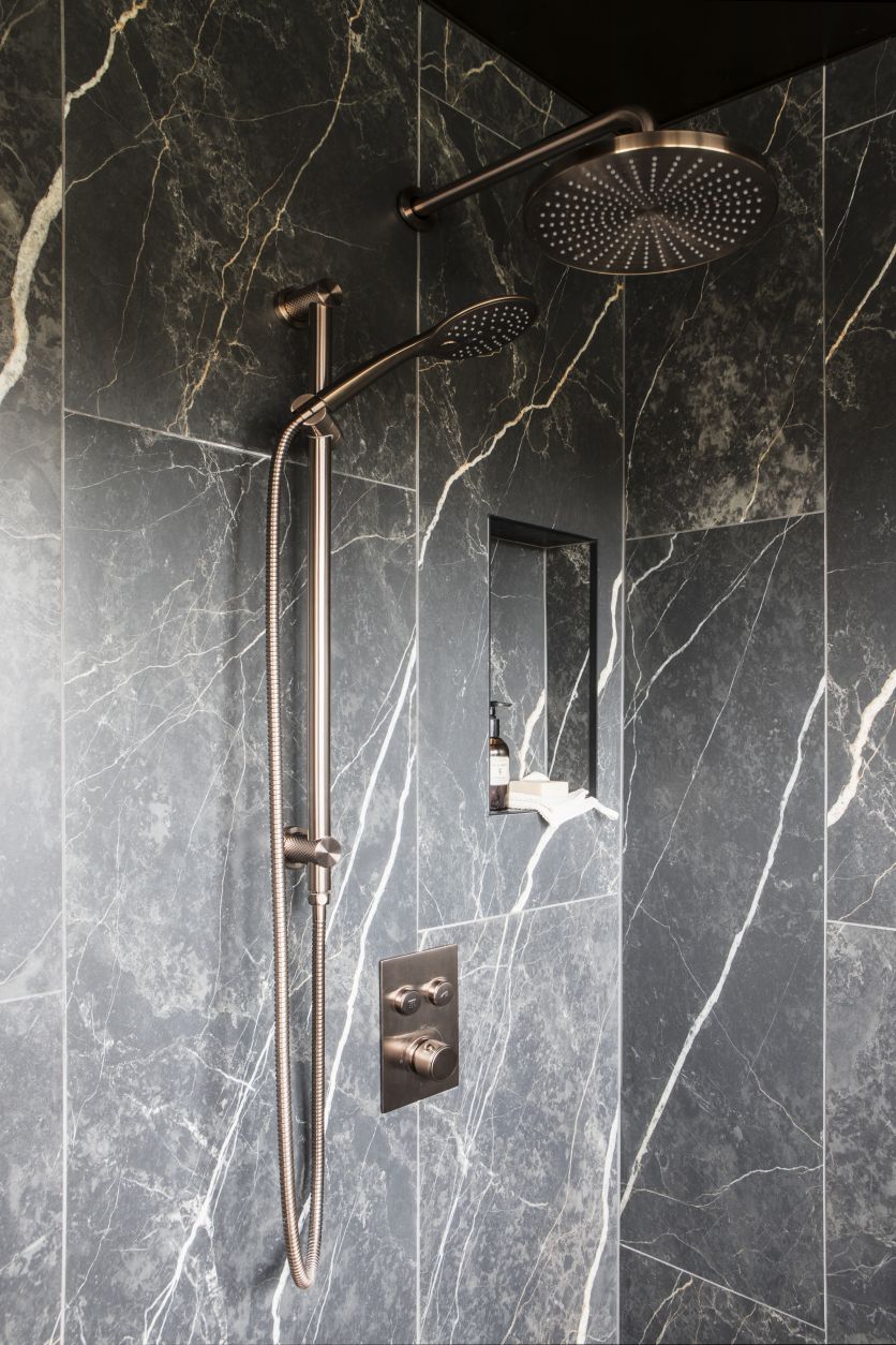 A luxurious walk-in shower featuring a large round rainfall head and separate handheld spray in a sophisticated brushed bronze finish, set against dramatic dark grey marble-effect wall tiles with striking white veining.