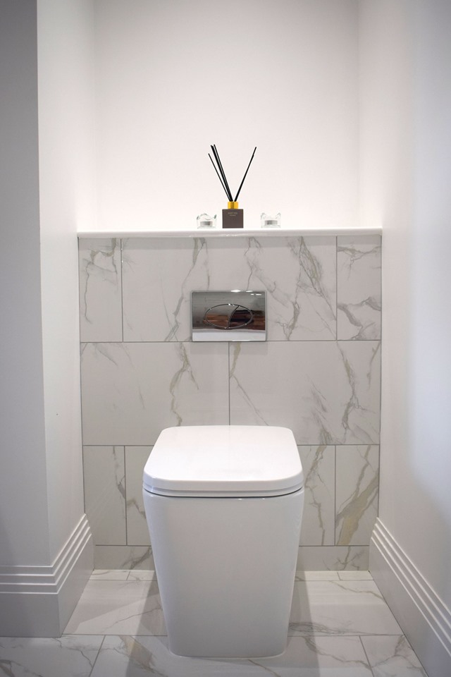 A contemporary back-to-wall toilet with a chrome dual-flush plate, set against elegant calacatta marble effect wall tiles in a minimalist white cloakroom.