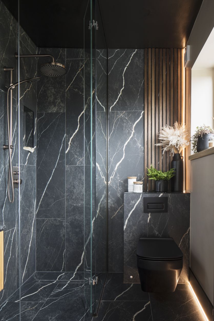 A luxury modern bathroom featuring dramatic black marble-effect wall and floor tiles, a walk-in glass shower with a brushed brass rainfall shower head, and a contemporary matt black wall-hung toilet set against a decorative wood slat feature wall.