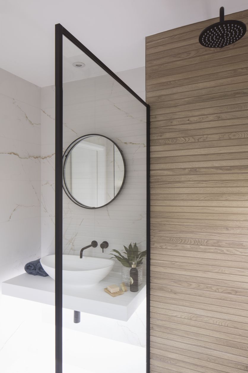 A luxury wet room featuring a black rainfall shower and framed glass screen, set against white gold-veined marble tiles and a textured wood-effect feature wall. A white countertop vessel basin with black wall-mounted taps completes the look.