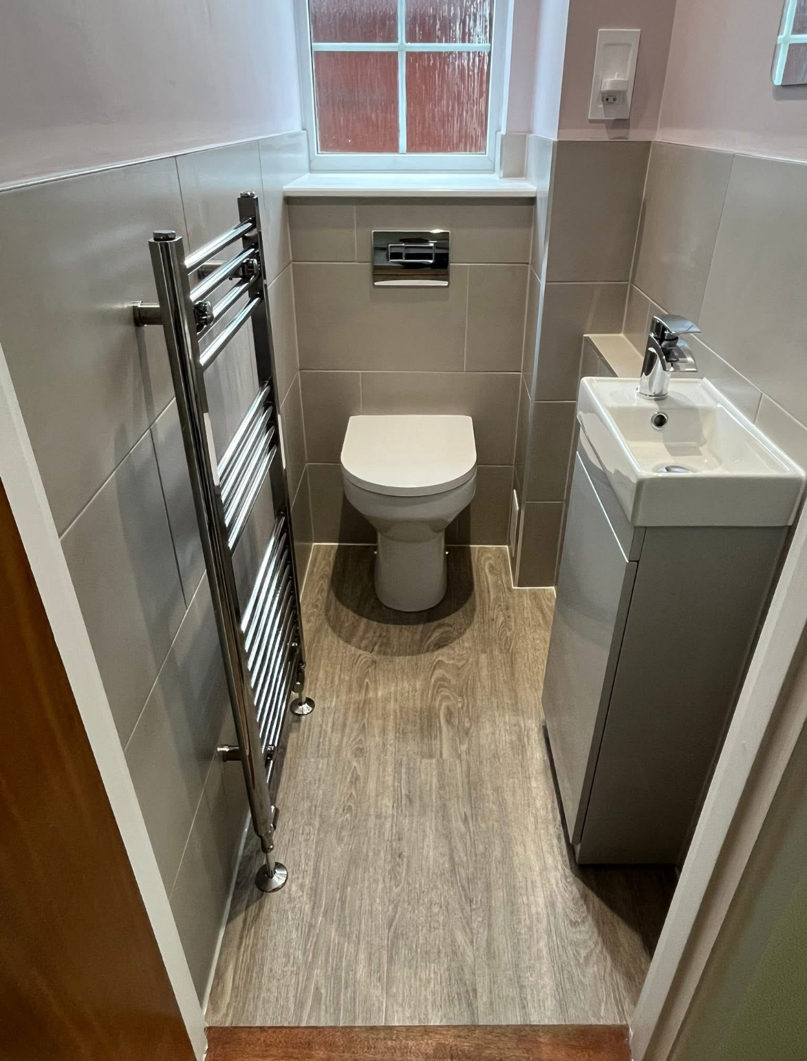 Completed compact cloakroom with a white toilet, vanity unit, dark radiator and wood-effect flooring.