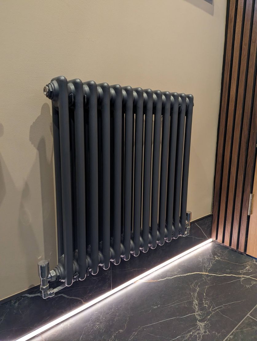 A horizontal anthracite three-column radiator with chrome valves mounted on a beige wall above dark marble floor tiles with integrated LED lighting.
