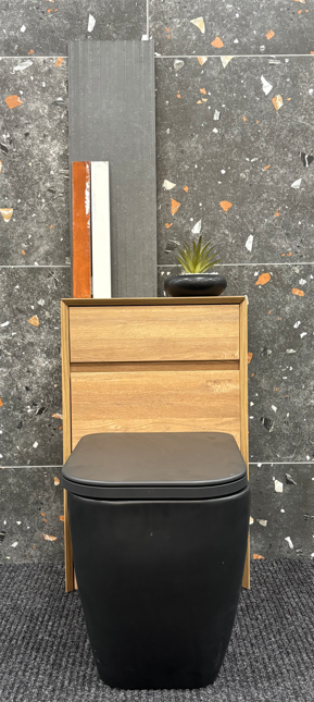 Compact bathroom display with a black toilet, wood-effect WC unit and dark terrazzo-style wall tiles with coloured flecks.