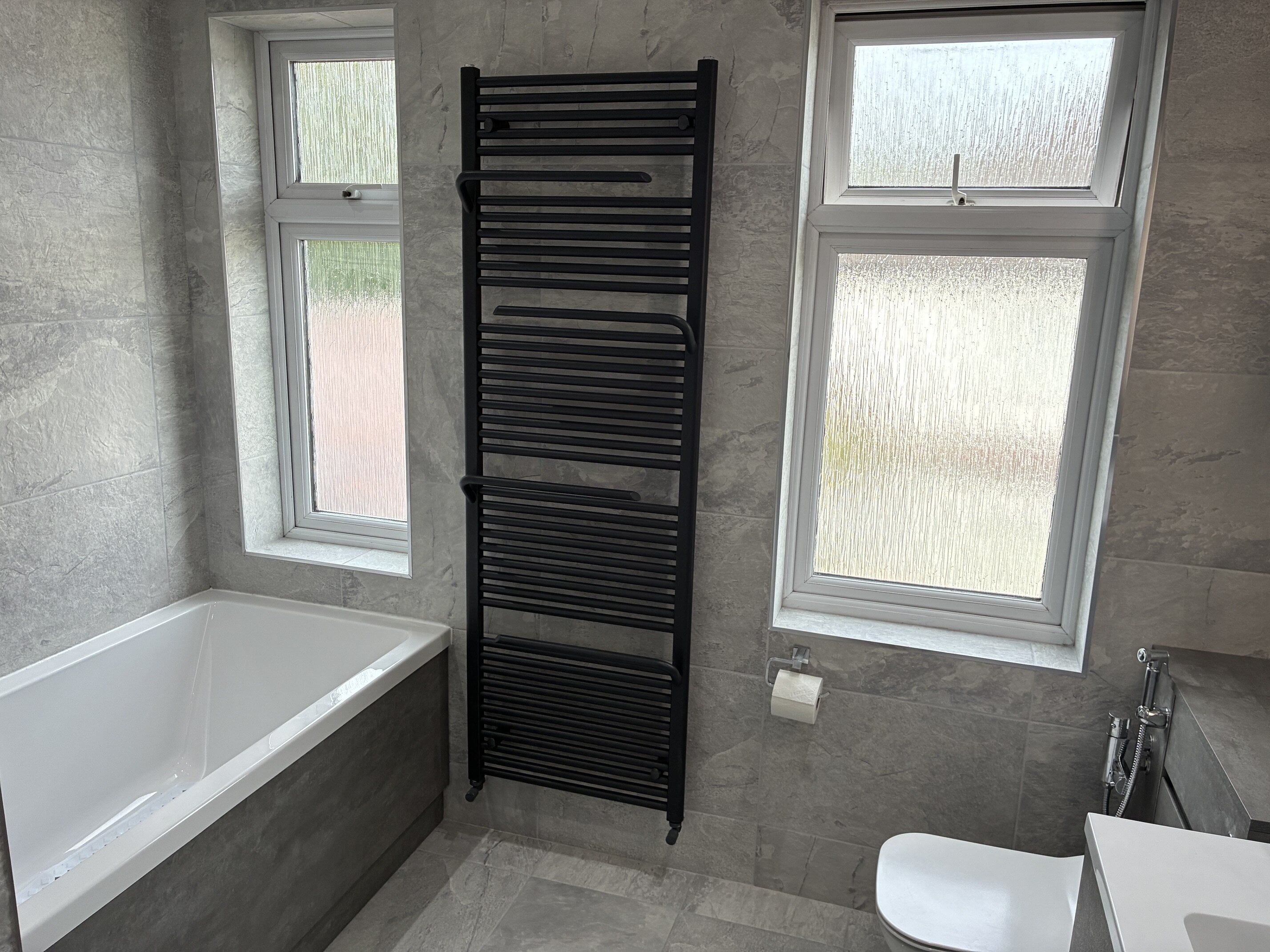 Completed bathroom featuring a fitted bath beneath two windows with grey wall tiles and a white toilet.