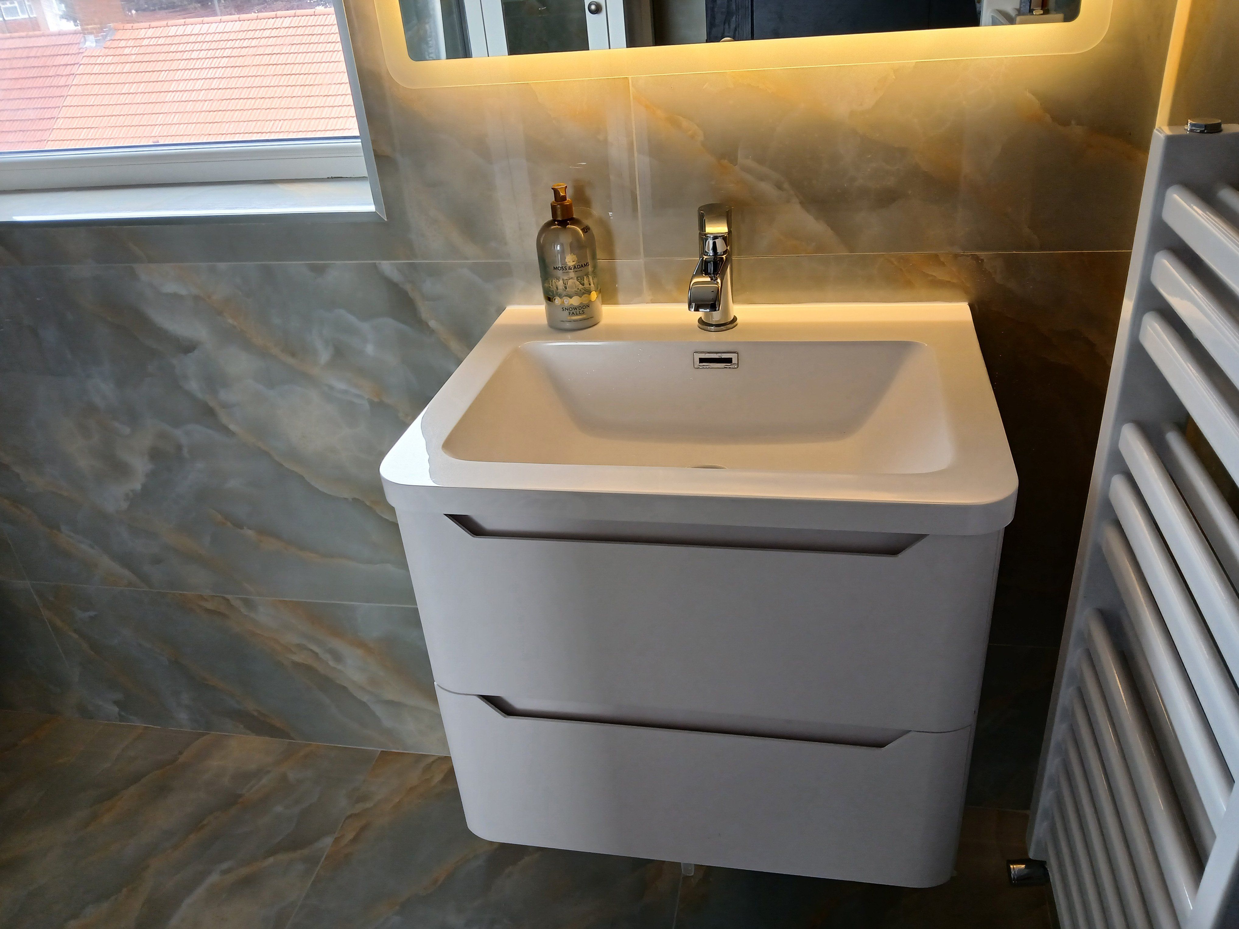 White wall-hung vanity unit with integrated basin, chrome tap and illuminated mirror against grey marble-effect tiles.