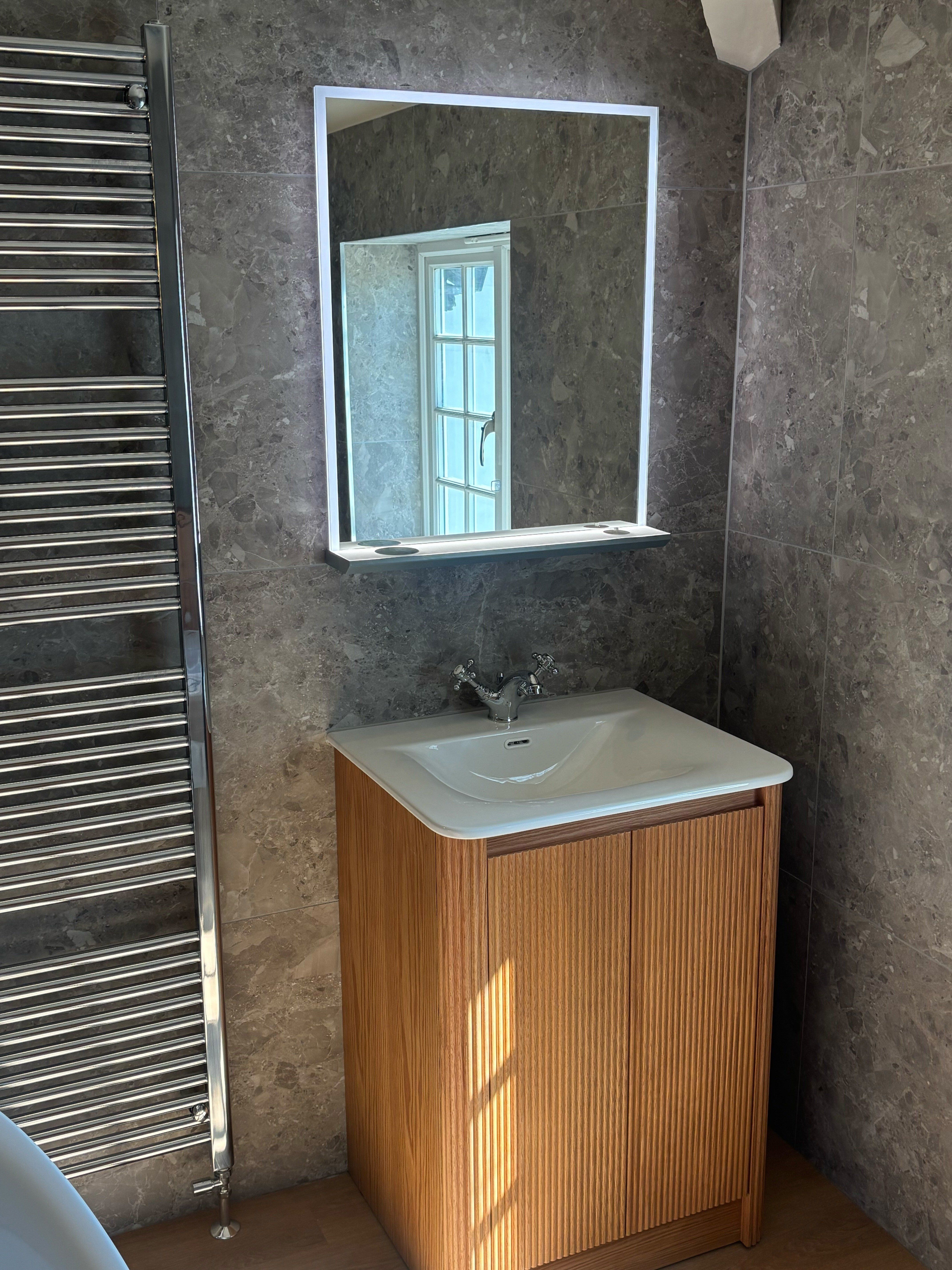 Natural oak bathroom vanity unit with white basin, illuminated mirror and neutral stone-effect wall tiles.