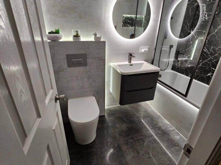 A modern industrial bathroom featuring Swindon white concrete-effect wall tiles, grey semi-polished floor tiles, and a La Bella black marble hexagon feature wall. It includes an Ellis charcoal vanity, Zena gunmetal taps, and a Felix backlit mirror.