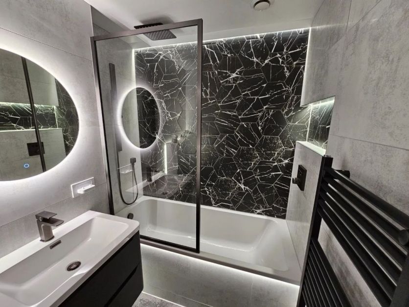 A monochrome bathroom featuring a La Bella black marble hexagon feature wall behind a bath, accented by an Amien gunmetal framed screen, Felix backlit mirror, and Swindon white concrete tiles.
