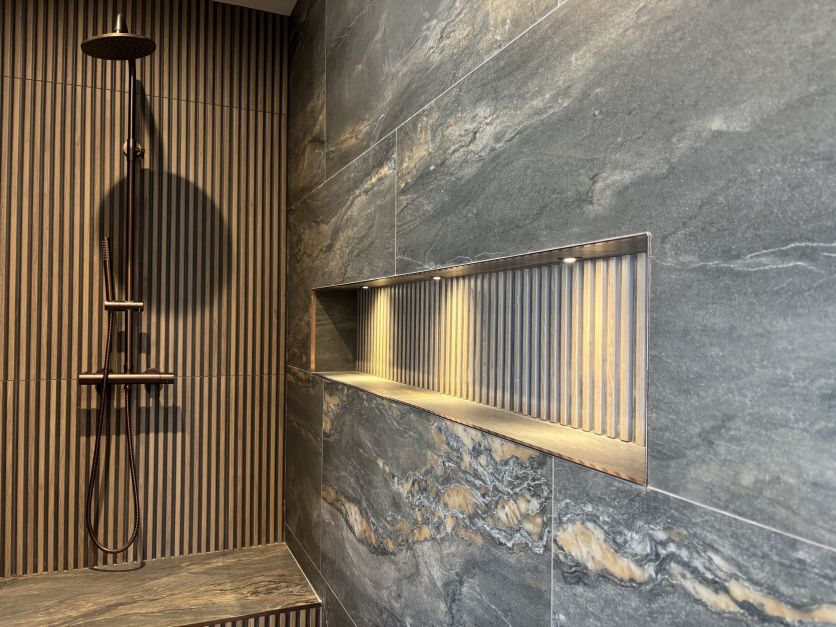 Luxurious brushed bronze shower and long illuminated niche set against elegant wood slat and dark veined stone tiles.
