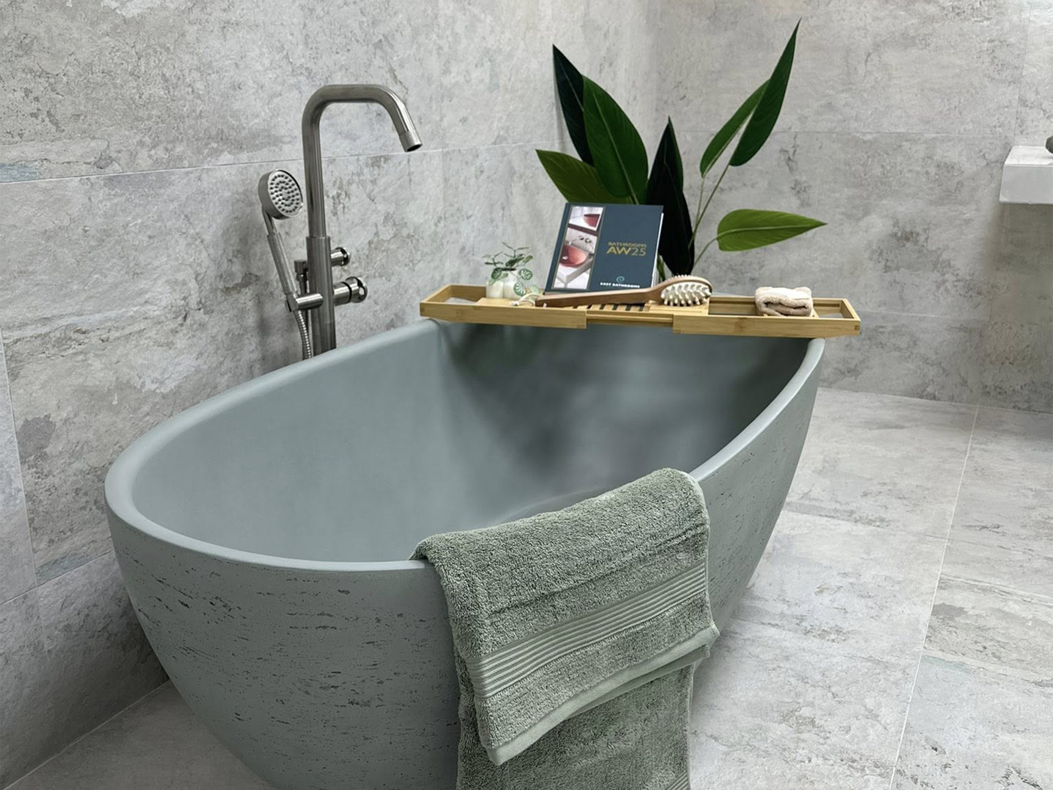 Grey freestanding bath styled with a bath tray, black tap, folded towel and plants in a modern tiled bathroom