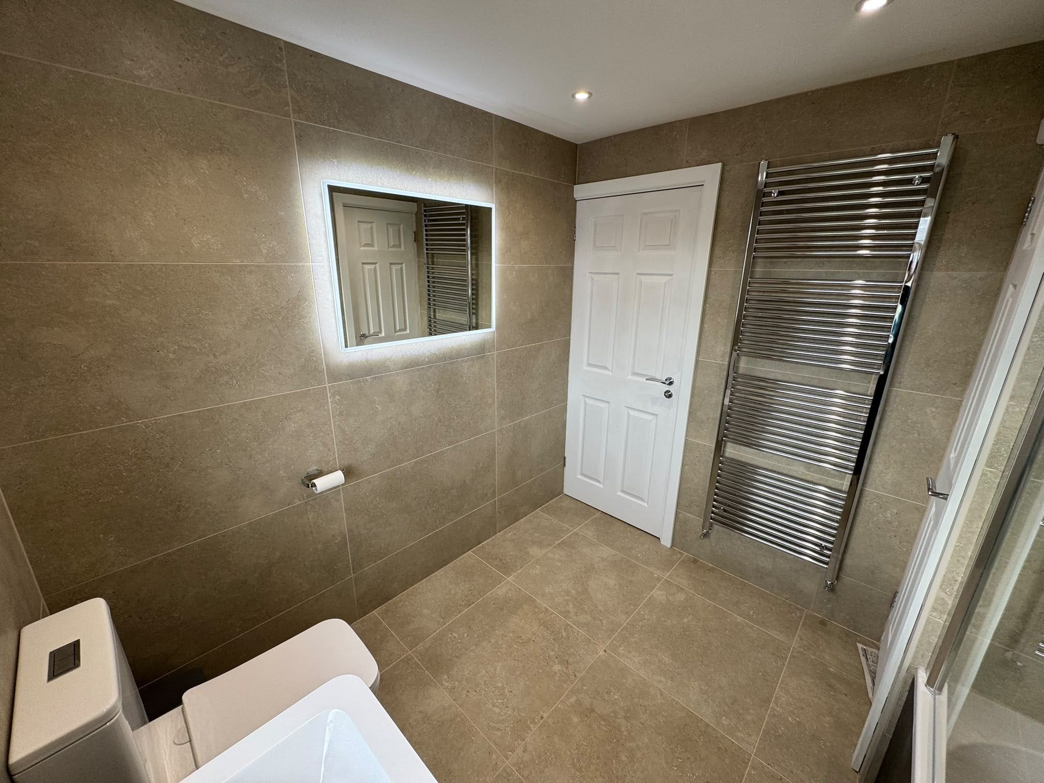Illuminated bathroom mirror above a white basin beside a chrome heated towel rail and glass shower enclosure.