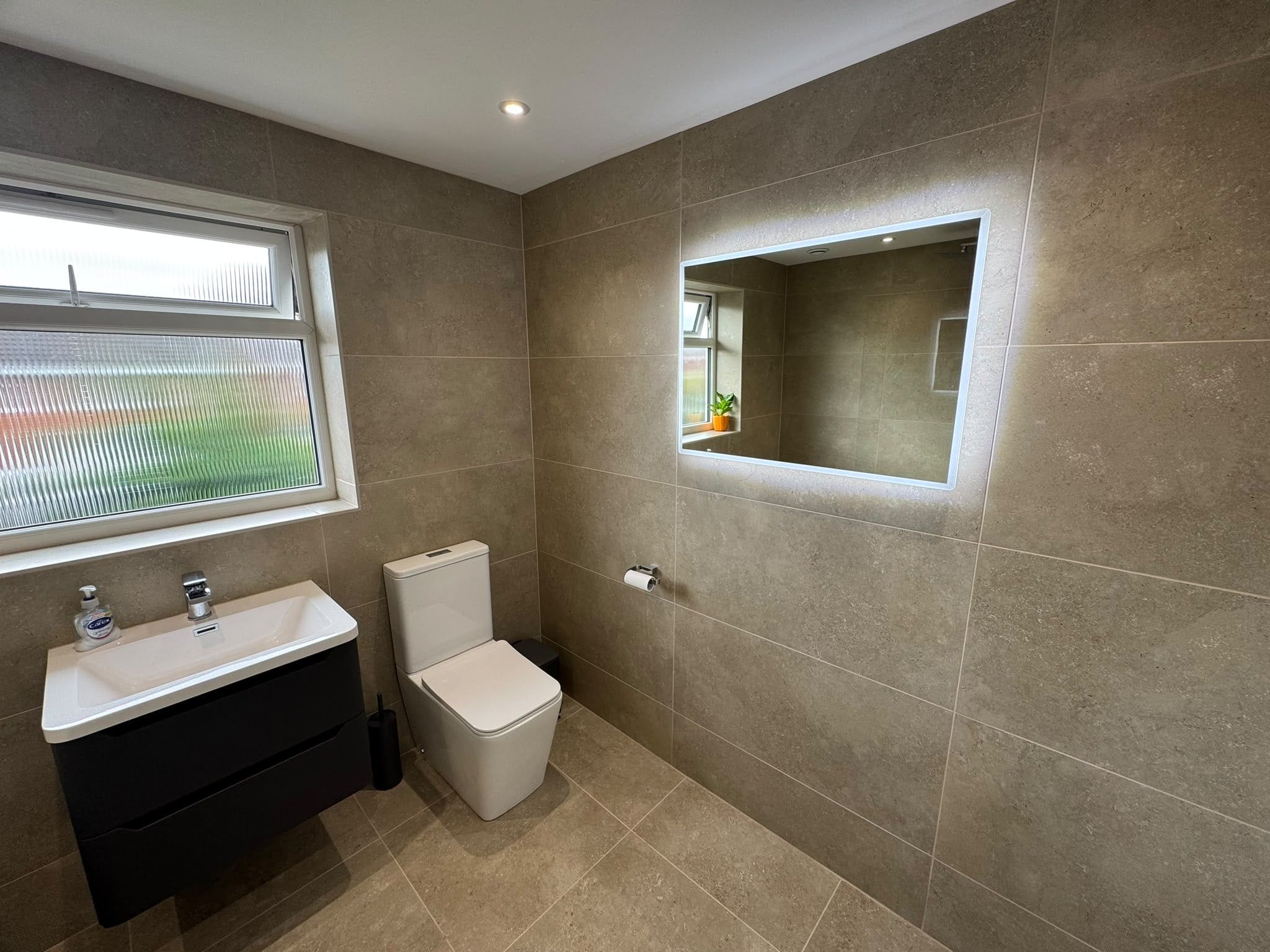 Matt black vanity unit with white basin and toilet beneath an illuminated mirror in a neutral tiled bathroom.